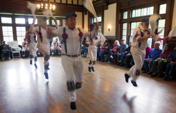 Six morris dancers with white shirts and trousers, blue caps, and grey suspenders decorated with red and blue ribbons, are in a large Tudor revival room. All are in motion, both hands coming up. Behind them is are some morris dancers in white. To the sides are audience members in chairs.