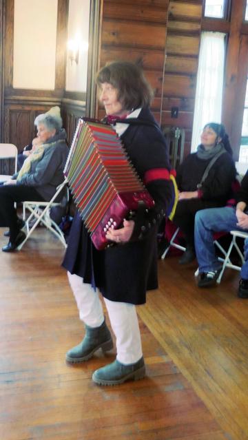 A musician in white shirt and trousers with a dark blue coat, decorated with red, blue, and yellow ribbons, in a wood paneled room, is playing a melodeon. Behind her are audience members in chairs.