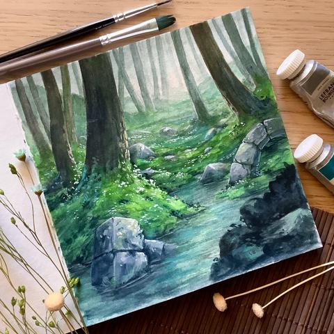 A photo of an open, square sketchbook placed on a wooden table, surrounded by paint brushes, paint tubes, and dried flowers for decoration. The sketchbook page features a few scattered chunks of an old stone ruin in a forest of smooth bare tree trunks. Moss and tiny white flowers cover the ground and part of a body of water forms the foreground. The lighting is hazy with a barely noticeable warm undertone.