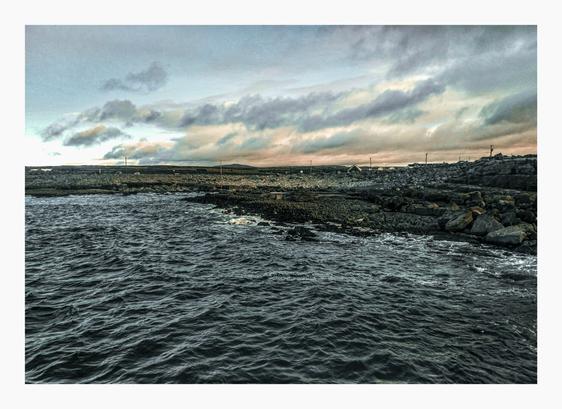 The photo is a seascape under a cloudy sky. In the foreground there is dark, choppy water. In the midground there is a rocky shoreline, with dark gray and black rocks meeting the sea - the rocks are closest to us on the right hand side and furthest away on the left. In the background there is aflat, sparsely populated landscape is visible under a sky filled with muted gray and pale pink clouds. There are some white coloured houses just visible in the distance.