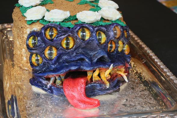 Scary purplish mimic face with 12 yellow eyes and a lolling red tongue that is on what looks like an otherwise normal birthday cake.