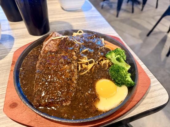 Sirloin steak, on a sizzling plate, smothered in black pepper sauce, egg frying on the plate, noodles, and some green and orange garnish.