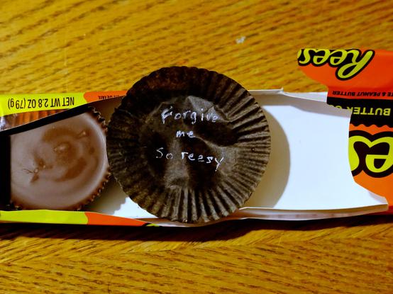 An opened package of Reese's Peanut Butter Cups. One on the left is still intact. On the right there is clearly an empty space where one or more cups used to be. In the center of the frame and the package is an empty paper wrapper from a peanut butter cup on which someone has written--in fine white paint or gel marker, no less--"Forgive me, So reesy."