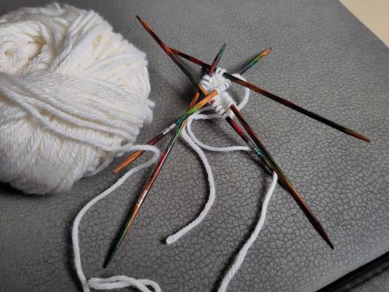 Picture of the beginning of a knitting project. On a grey background, a ball of white yarn, and a sort of mikado made of 5 double-pointed needles held together by the first rows of a sock.
