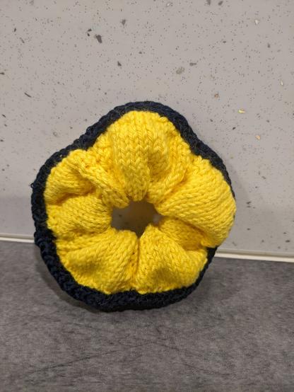 A knitted scrunchie. It is yellow with a dark blue hem surrounding it.