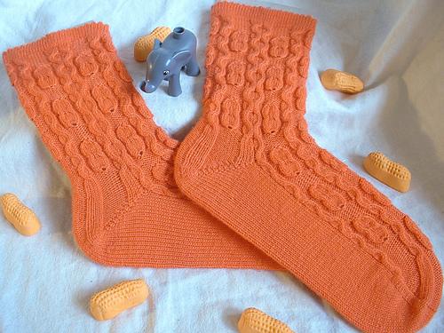 A cozy pair of bright orange socks with a pattern reminiscent of peanuts laid out flat, there is a small toy elephant between them, and they are surrounded by a scattering of orange candy circus peanuts.