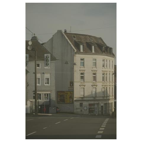 Blick über eine leere Straße hinab auf einen Straßenzug in der Morgensonne.  Kein Auto ist zu sehen. Einsam hängen Ampeln über der Straße.