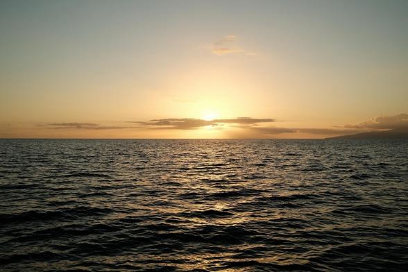 a boat view, but with no boat visible, of a sunset over the Pacific ocean with a 98% clear sky and gentle waves, creating a serene and peaceful atmosphere. To the right, an island can be seen on the horizon. It is either Maui, Moloka’i, or Lana’i