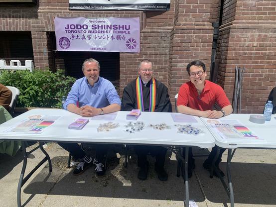 Three men sit at a table covered in pins, pamphlets, and Pride posters. Two are in regular street cloths and one is wearing a black ministerial robe, with a rainbow wagesa (stole-like abbreviated robe). Behind them a lilac, purple, and white sign reads "Jodo Shinshu Toronto Buddhist Temple," in both English and Japanese.