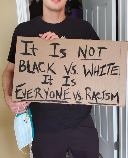 A man in black holding a cardboard sign that reads "It is not black vs white - It is everyone vs racism"

He's holding a mask 😷 because of covid.