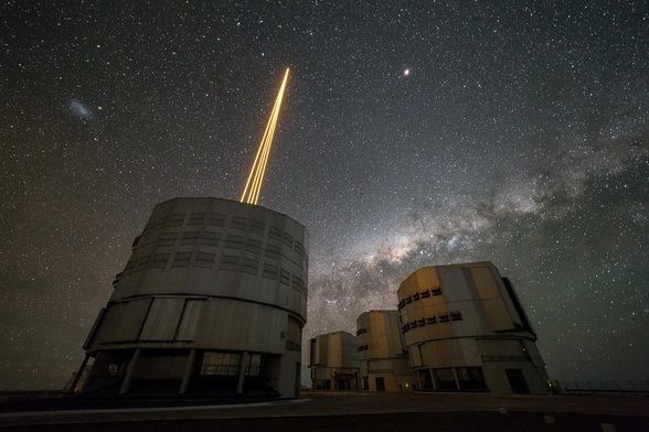 A night photograph showing four large cylindrical telescope domes under a dark starry sky. One of them is firing four yellow laser beams that appear to converge on one point in the sky. The bright band of the Milky Way crosses the lower part of the sky.