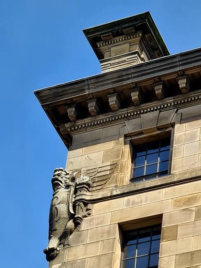 A winged griffin-like creature with a webbed foot holding a cartouche on the corner of a 1920s sandstone building.
