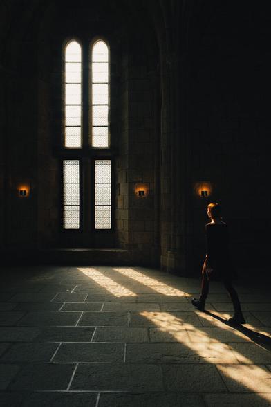 A woman walks towards a big church window, following the sunbeam the window casts. Three orange lamps match the woman's illuminated hair.