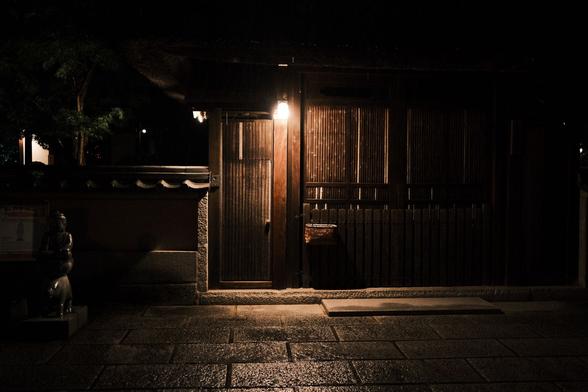 A lamp lights a doorway of a traditional home in Kyoto’s Gion district. The building is wood. Stone pavers are wet with rain in the foreground.