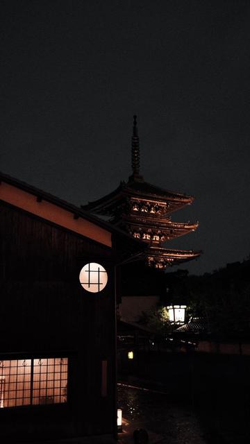 A tall pagoda rises in the night behind a traditional building in Kyoto’s Gion district. The building in the foreground has circular and rectangular windows that are lit from within. A streetlamp lights the crowded, wet streets.