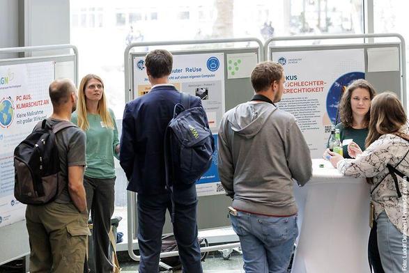 Gruppe von Menschen während einer Ausstellung mit Informationspostern im Hintergrund, einige diskutieren an einem Tisch.