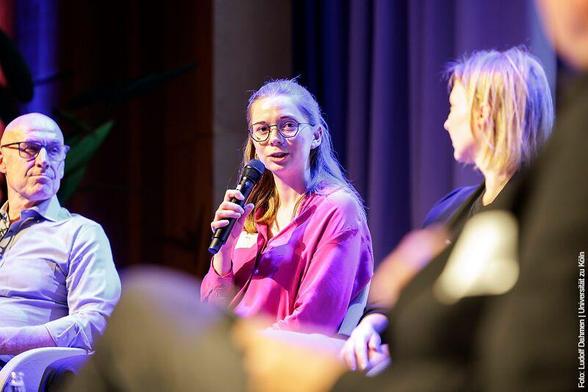 Person im pinken Hemd spricht in ein Mikrofon bei einer Podiumsdiskussion, begleitet von zwei weiteren Personen sitzen rechts und links.