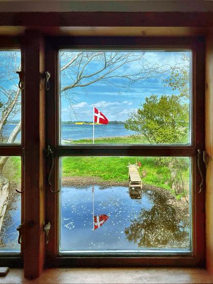 Blick aus einem Sprossenfenster aufs Meer, gegenüber das Festland, im Vordergrund ein Teich mit Steg vor einer gehissten dänischen Fahne neben Bäumen vor einem blauen Himmel.

View from a mulleted window to the sea, opposite the mainland, in the foreground a pond with a footbridge in front of a hoisted Danish flag next to trees in front of a blue sky.