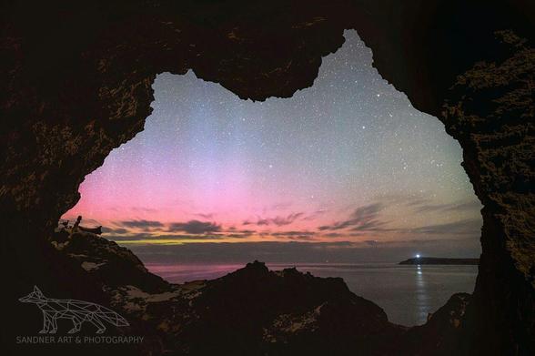 A vibrant night sky viewed through a cave opening, shaped uncannily like the outline of mainland Australia. The aurora australis glows across the southern sky in soft bands of pink, purple, and green above a calm ocean. A lighthouse glows faintly in the distance, casting a narrow reflection on the water. The cave walls are silhouetted, framing the scene in deep contrast and guiding the eye toward the glowing horizon and star-filled sky.