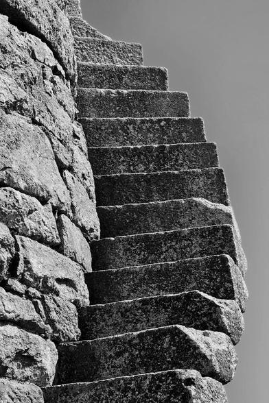 A low-angle, black and white shot of rugged stone steps ascending around the textured stone wall of an old furnace chimney, silhouetted against a plain, light grey sky. The steps appear ancient and worn, leading the eye upwards.
