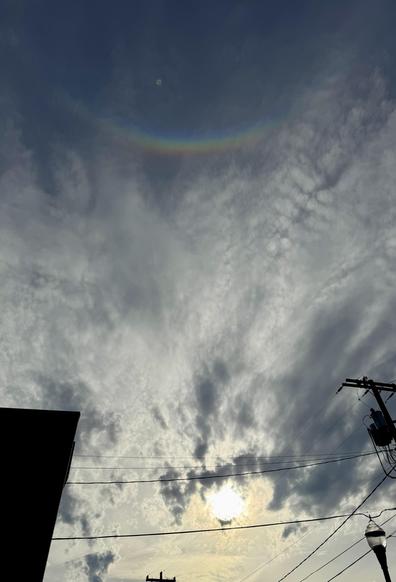 The image features a cloudy sky with streaks of light and a faint rainbow arc near the top. The sun is partially obscured by the clouds, and silhouettes of buildings and utility poles are visible in the foreground.