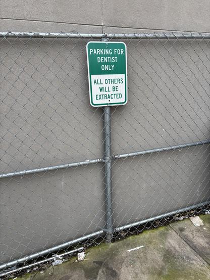Grey wall with chain link fence directly in front of it. A green and white sign says “Parking for Dentist Only - All Others Will Be Extracted”. Some mossy cement is seen at the base of the fence.