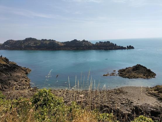 Photographie d’un paysage marin sous un ciel clair et légèrement voilé. À l’avant-plan, des herbes blondes et des rochers sombres bordent une petite crique. L’eau turquoise s’étale paisiblement jusqu’à un îlot rocheux allongé, qui s’élève au centre de l’image comme un dragon endormi. À droite, un minuscule kayak turquoise fend doucement la mer, ajoutant une touche de solitude libre et d’aventure à cette scène sauvage et magnifique.