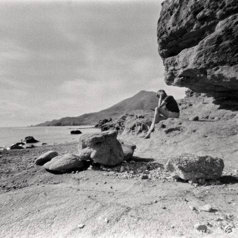 A black and white pinhole photograph of a lady sat on a rock and reading a book in a secluded bay by the sea