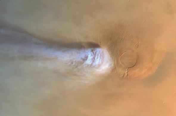 Color image of Mars featuring Arsia Mons: a tan, dome shaped shield volcano with a broad circular caldera and subtle concentric ridges at right. A bright whitish elongated cloud emerges from the volcano’s western flank and stretches horizontally leftward, fading across a hazy atmosphere.