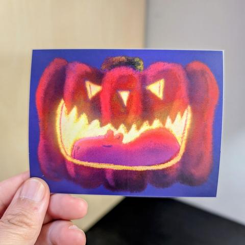 Photo of a left hand holding a rectangular sticker. The sticker is a matte painting of a capybara sleeping in a glowing jack o lantern.