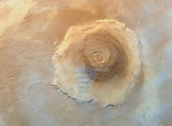 An image from orbit taken by the ESA Mars Express spacecraft shows Olympus Mons, one of the tallest volcanoes on Mars. Its massive, circular shield-like structure dominates the center, appearing as a broad, raised dome with a gently sloping profile. At the summit lies a large, irregularly shaped caldera with several circular pits resembling overlapping depressions. Concentric bands of darker and lighter tones surround the caldera. Steep cliffs mark the volcano’s outer edge. The surrounding Martian surface displays varied textures: to the left, wrinkled, ridged plains resemble frozen waves, to the right and lower portions, the terrain smooths out. The colour palette includes browns and rust-like reds, with a hint of whitish cloud. Lighting from the upper right casts soft shadows, enhancing the volcano’s three-dimensional form and surrounding landscape.