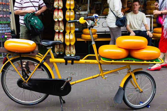 In the foreground, photographed from the side, a yellow bicycle (over the entire length of the photo) with a large round yellow cheese on the luggage rack on the back (left) and three large round yellow cheeses stacked on top of each other on a luggage rack in front. In the background, a cheese shop in The Hague. On the left, a door with a vertical row of yellow clogs to the right of it. In front of the window, a bench with more cheeses and a seated man. Here and there, a few more people, but they are only half visible. The bell of the bicycle is also a small cheese.