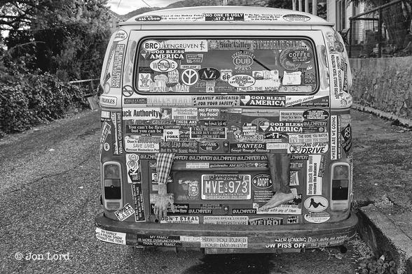 This Is A Black And White Street / Humorous Photo In Landscape Format. Offset To The Right Is The Rear Of A VW Microbus (Officially A Volkswagen Type 2). The Image Is Aligned So That The  Back Is The Only Part In View. The Rear Of The Micro Bus Is Flat Not Streamlined. The Surface Is Covered By Hundreds Of 'Bumper Stickers'. Examples Are 'Honk If You've Has Sex With The President' (Clinton Era). 'My Family Is More Dysfunctional Than Yours' And 'I'll Try Being Nicer If You Try Being Smarter'. The Microbus Is Parked On A Narrow Street With Unkept Vegetation On The Left Side, A Pavement (Sidewalk) On The Right With A Brick Wall And A Partial View Of A Residential House Beyond.
The Location Jerome, Arizona (2016).