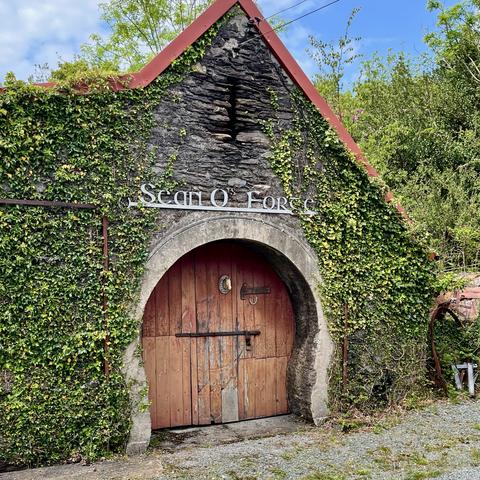An ivy covered single story fieldstone building. A wooded door with an iron latch sits in an opening that starts smaller at the bottom, then sweeps out towards the rounded top.