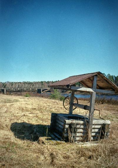 In the foreground is an old, wooden well in the middle of sand-colored dry grass. A bridge, part of the river and the forest can be seen in the far background.