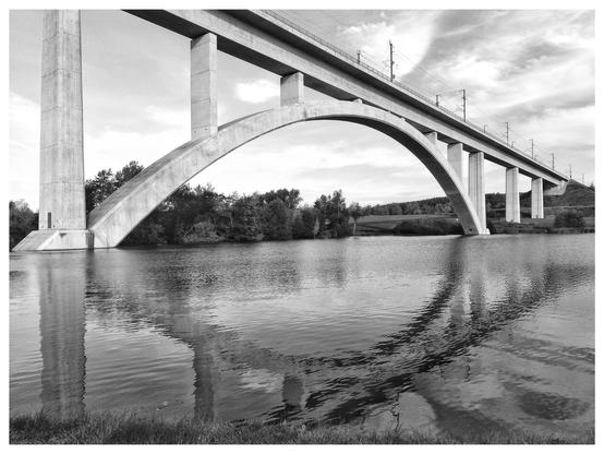 Schwarz-Weiß-Foto mit weißem Rand. In einem Teich steht eine Betonbrücke für den ICE-Verkehr, sie spiegelt sich im Wasser.