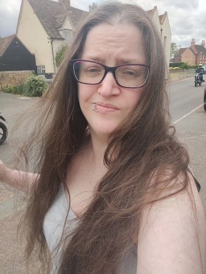 A photo of me walking back from the postbox. I'm a white person with long brown hair, glasses, and a lip ring. I'm squinting a bit because natural light burns (even though it's very overcast). I'm wearing a gray tank top, and you can see a house across the road behind me.