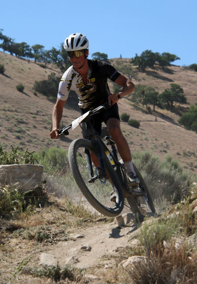 Our youngest son in his last race of his high school career, Tehachapi, 5/25/2025. He is going over the final rocky section before turning down to the finish line. His front tire is up in the air and a cloud of dust is behind him. In this race he got a flat tire on the first lap, fixed it, and made up all of the positions he lost. The hills are dry behind him. The skies are blue, but it was windy.