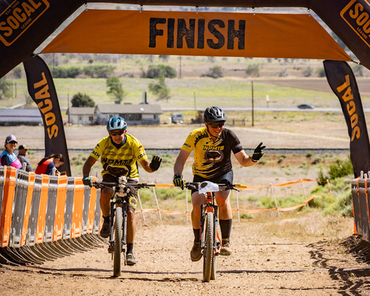 Two Newbury Park High School mountain bike team coaches, in their yellow coach jerseys, cross the finish line in the Coach race at the league Championships weekend in Tehachapi, CA, 5/24/2025. This was a fun race to let the coaches compete on the "middle school" course. Coaches created their own race plates. Mine was a stuffed kitty (I'm the rider on the left).