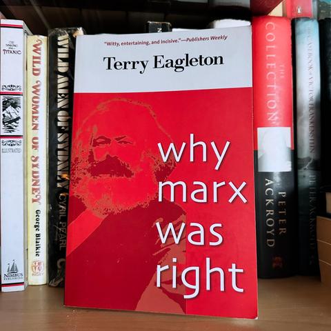 The image features the book "Why Marx Was Right" by Terry Eagleton, displayed on a bookshelf. The book cover is predominantly red, featuring a depiction of Karl Marx. Several other books are visible in the background, including titles related to history.