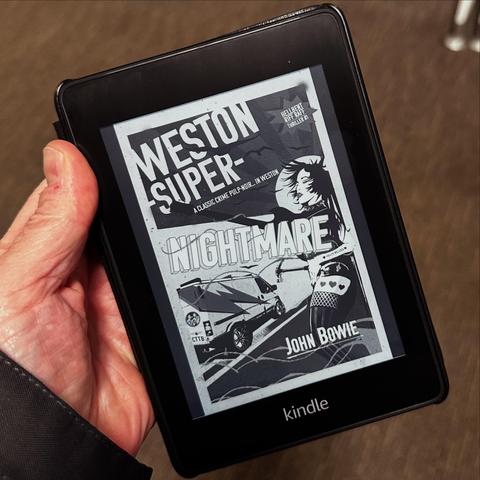 An e-reader displaying the cover of the book "Weston Super Nightmare" by John Bowie. The cover features a vintage design, showcasing elements of crime and noir, with the title prominently displayed. A hand is holding the device, with a blurred background.