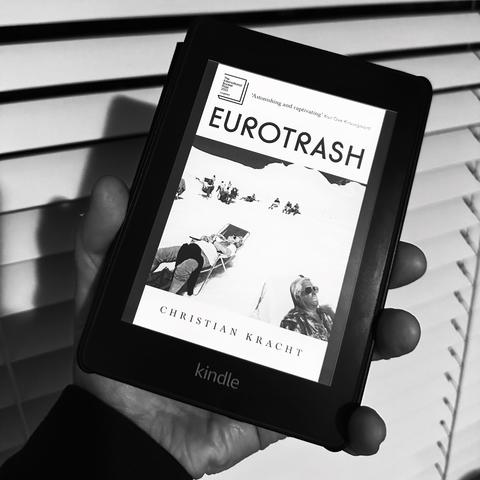 A hand holding a Kindle displaying the cover of the book "Eurotrash" by Christian Kracht. The cover features a black and white image of people relaxing on snow, with the title prominently displayed at the top. Blinds are in the background.