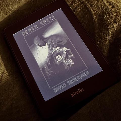 An e-reader displaying the cover of the book "Death Spell" by David Sodergren. The cover features dark, artistic imagery, including a skull with an eye and abstract elements. The device is resting on a textured surface.