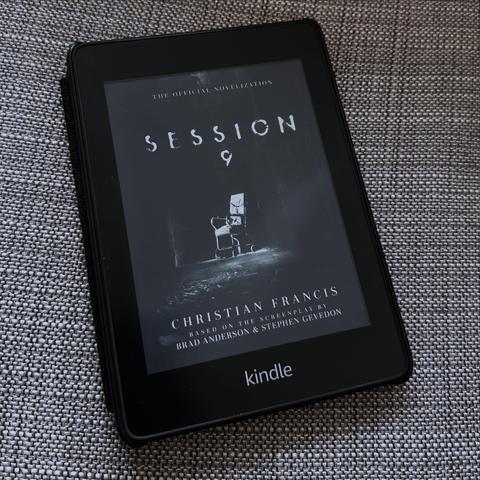An e-reader displays the cover of "Session 9," the official novelization by Christian Francis, based on the screenplay by Brad Anderson and Stephen Gevedon. The cover features a dark, eerie scene with an empty chair in a dimly lit hall.