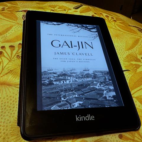 An e-reader displaying the book "Gai-Jin" by James Clavell. The cover features a historical black-and-white image of a Japanese town with ships in the background. The device is resting on a patterned yellow surface.