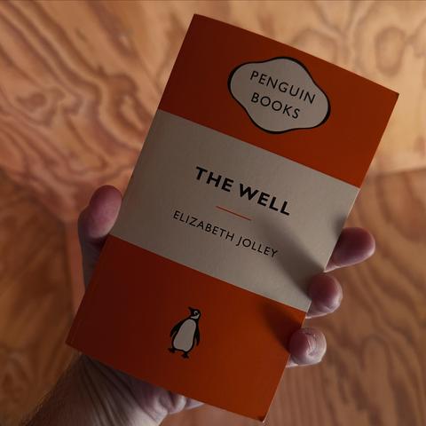 A hand holds the book "The Well" by Elizabeth Jolley, published by Penguin Books. The book features an orange cover with a white band and the Penguin logo at the bottom. The background has a warm, wooden texture.