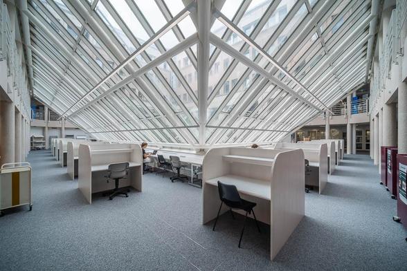 Image of a spacious, modern library. Rows of individual study cubicles with chairs are organized under the angled glass roof. A person is seated at one of the desks.