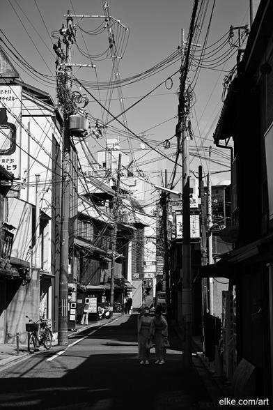 A striking black-and-white street scene from Kyoto, Japan, capturing the visual chaos of overhead power lines that crisscross the sky like a web of modern life.

In the foreground, two women in traditional kimonos walk through the quiet alleyway, blending old and new Japan in a powerful contrast. The strong architectural lines, urban textures, and delicate human presence create a compelling moment of stillness in a dense cityscape.

Perfect for minimalist interiors, modern hallways, or monochrome-themed rooms.

Ideal for collectors of Japanese urban photography or lovers of street scenes that tell layered stories.