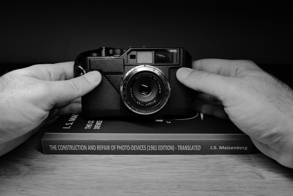 A digital monochrome photo showing a pair of hands holding a Voigtländer Bessa R2C rangefinder camera, fitted with a Carl Zeiss 35mm f/2.8 Biogon lens in Contax RF mount. The camera rests on a hardcover book titled “The Construction and Repair of Photo-Devices (1961 Edition) – Translated” by I.S. Maizenberg. (Photo by me, CC-BY-4.0)