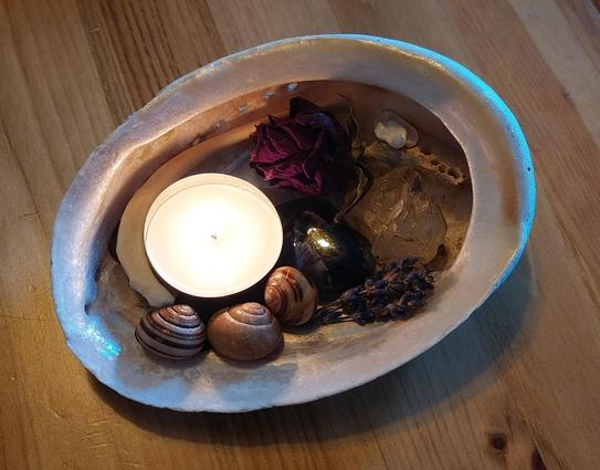 A lit tea light in a large shell with bits of dried lavender, a dried red rose head, some snail shells, stones, and bones, on a wooden table.